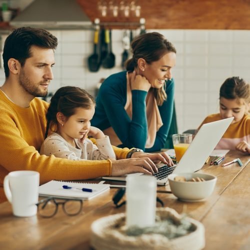Working father using computer while daughter is sitting on his laptop at home. Mother and daughter are doing homework in the background.